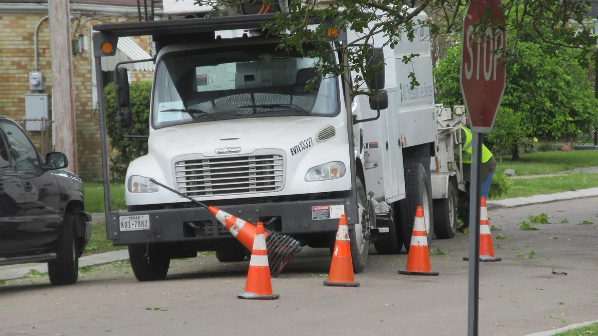A white bucket truck parked curbside behind orange cones on a tree-lined street.