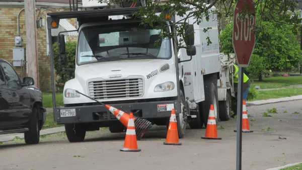 A white bucket truck parked curbside behind orange cones on a tree-lined street.