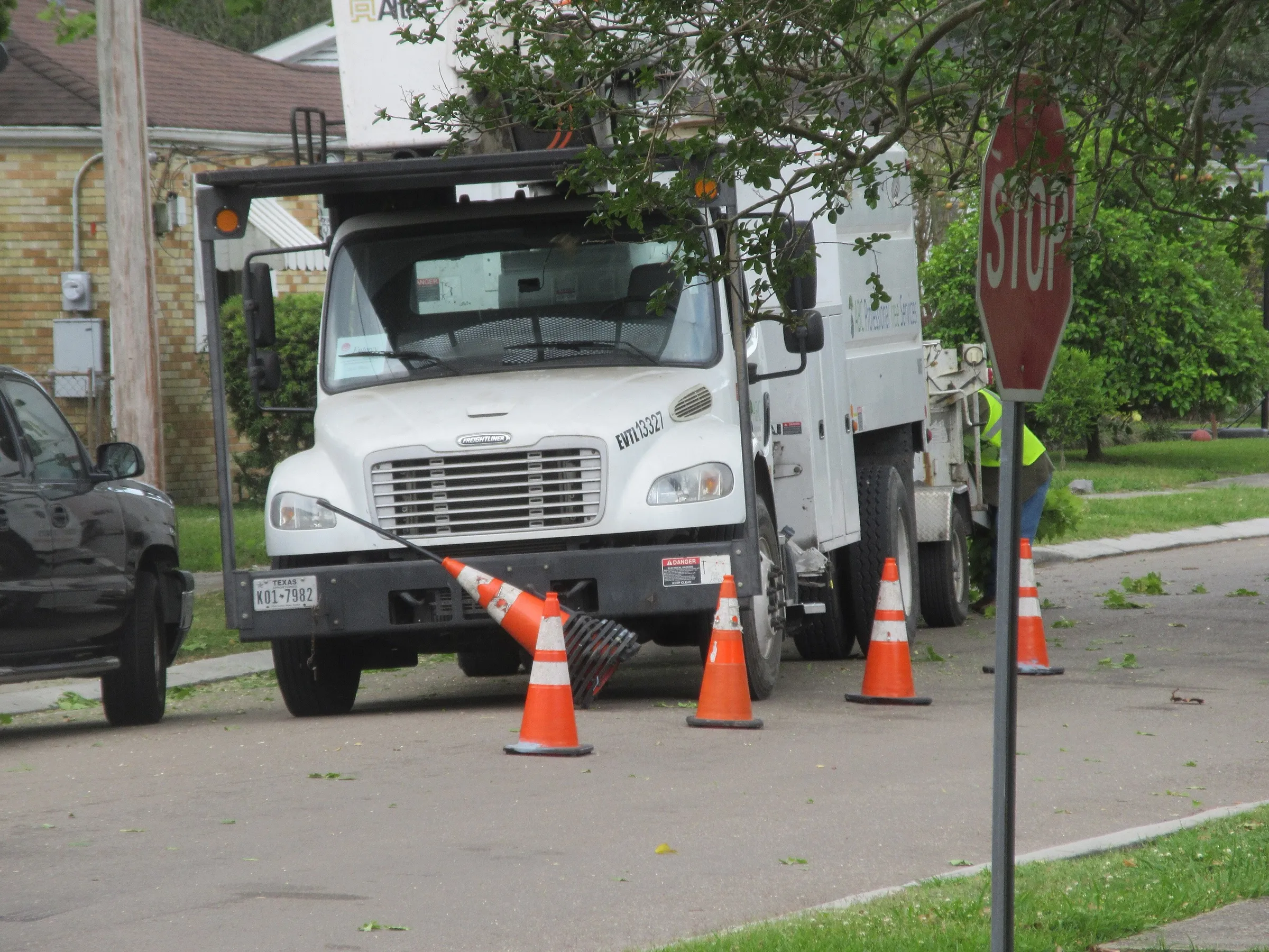 A white bucket truck parked curbside behind orange safety cones on a tree-lined street.