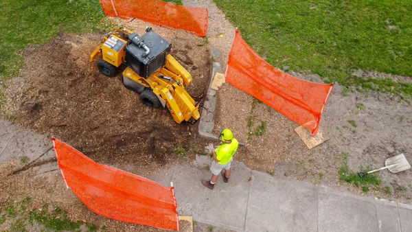 A compact stump grinder working inside a fenced-off patch of lawn beside a sidewalk.