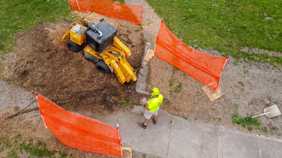 A compact stump grinder working inside a fenced-off patch of lawn beside a sidewalk.